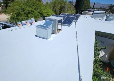 A white-painted flat rooftop with solar panels, an air conditioning unit, and paint supplies. Trees, a yard, and mountains are visible in the background under a clear blue sky.