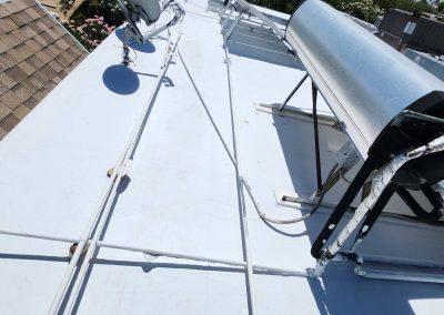 A white rooftop with a large solar water heater, satellite dishes, and cables running across the surface. There are parts of nearby buildings and greenery visible in the background under a clear sky.