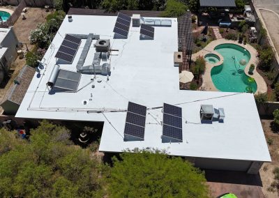 Aerial view of a flat-roofed house with multiple solar panels installed, surrounded by trees and yards. There is a kidney-shaped swimming pool and patio area in the backyard.