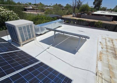 A flat white rooftop with solar panels, an air conditioning unit, and metal ductwork. There are trees and neighboring buildings in the background under a clear blue sky.