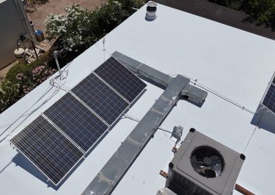 A rooftop with multiple solar panels, a ventilation unit with visible fan, and ductwork. Surrounding greenery and parts of neighboring roofs are also visible.