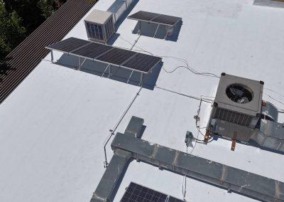 A rooftop with solar panels, air conditioning units, metal ductwork, and electrical wiring; trees are visible along the left edge of the roof.