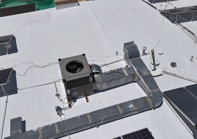 Aerial view of a flat white rooftop with solar panels, an HVAC unit, ductwork, and various pipes and vents; a swimming pool is visible in the background.