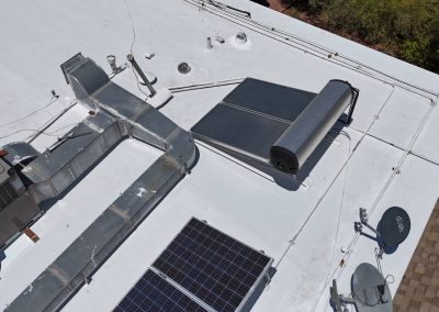 A white flat rooftop with solar panels, a solar water heater, HVAC ductwork, an air conditioning unit, and two satellite dishes, surrounded by trees and shingles at the roof’s edge.