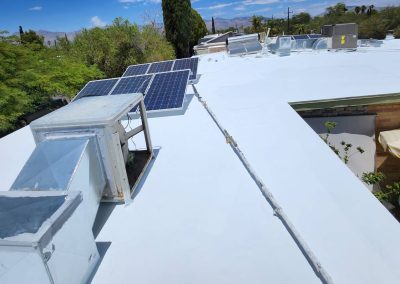 A white flat rooftop with solar panels, HVAC units, and ducts. Trees and a few rooftops are visible in the background under a blue sky with scattered clouds.