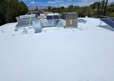 A white flat rooftop with solar panels, an air conditioning unit, and various vents under a blue sky with scattered clouds. Trees and distant mountains are visible in the background.