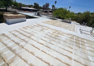 A flat white rooftop with patches of worn or faded coating, air conditioning units, a potted plant, and a ladder along the edge; trees and a residential street visible in the background under a clear sky.