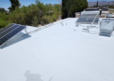 Flat white roof with mounted solar panels, a water heater, and various rooftop fixtures; trees and distant mountains are visible under a clear blue sky.