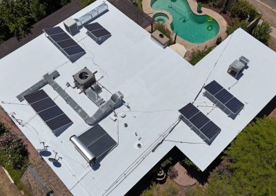 Aerial view of a flat white rooftop with multiple solar panels installed, various HVAC units, and a nearby backyard pool surrounded by greenery and a fence.