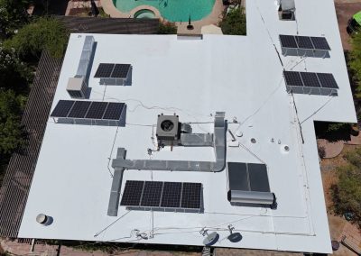 Aerial view of a flat white rooftop with multiple solar panels installed, ventilation units, and wires visible; a swimming pool and greenery are seen in the background.