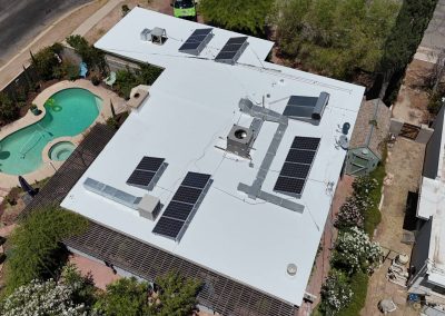 Aerial view of a flat-roofed building with multiple solar panels installed on top, surrounded by trees, a fenced yard, a swimming pool, and nearby roads.
