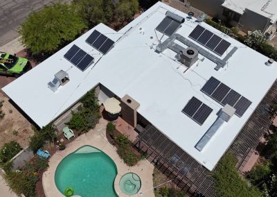 Aerial view of a white-roofed house with multiple solar panels, a pool, and circular hot tub in the backyard. There are plants, outdoor furniture, and a green utility vehicle parked on the street nearby.