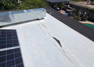 A flat white rooftop with solar panels and a large crack running through the surface. Nearby, there are metal structures, greenery, and a glimpse of a swimming pool and yard below.