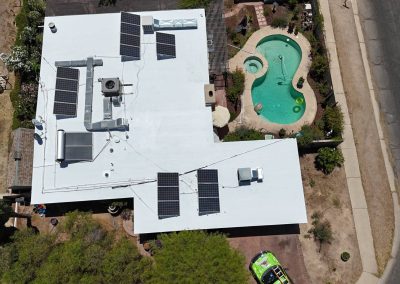 Aerial view of a white flat-roofed house with solar panels, a kidney-shaped backyard pool, surrounding patio, plants, and a green car parked nearby, next to a residential street.