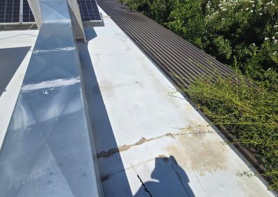 A rooftop with solar panels, a metallic duct, cracks and stains on the white surface, and green bushes along the edge. A person's shadow is visible in the foreground.