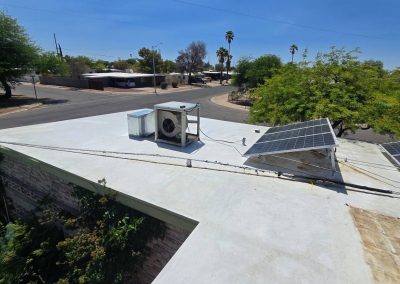 A white flat rooftop with two solar panels angled toward the sun and a large air conditioning unit. Trees surround the building, and a quiet suburban street with houses and palm trees is visible in the background.