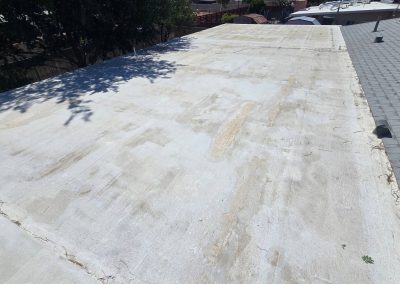 A flat, light-colored rooftop with uneven patches and some debris is shown next to a gray shingled roof. Trees and neighboring buildings are visible in the background.