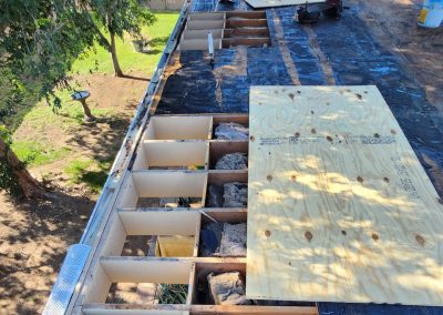 Roof under repair with several sections of plywood removed, exposing wooden beams and insulation. A new plywood sheet is partially installed. Trees and a grassy yard are visible beside the house.