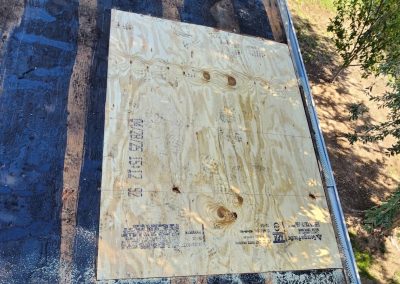 A piece of plywood has been installed on part of a roof. The surrounding roof area is covered in dark, weathered material. Trees and sunlight are visible to the right.