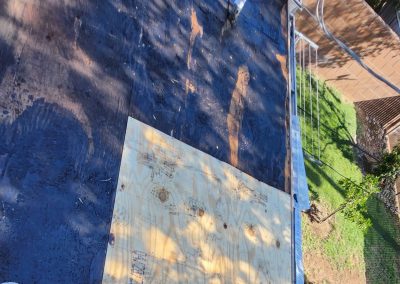 View of a roof under repair, showing a partially installed sheet of plywood on the dark roof surface. Some tools and safety railing are visible, with a green yard and a brown fence below. Shadows from trees cover parts of the scene.