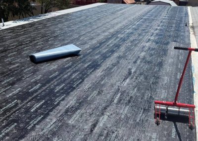 A flat roof covered with black roofing underlayment, a roll of material, a red roller tool, and a gray cap laying on the surface. Part of a shingled roof and tree branches are visible in the background.