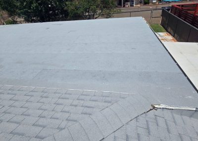 A rooftop view showing the transition between a shingled roof in the foreground and a flat, gray, asphalt roof in the background, with trees, a street, and fencing visible beyond the roof.