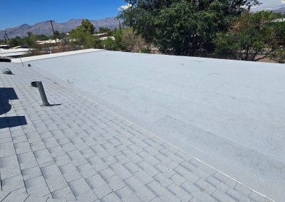 View of a flat, light gray roof next to an angled shingle roof, with trees and mountains in the background under a clear blue sky.