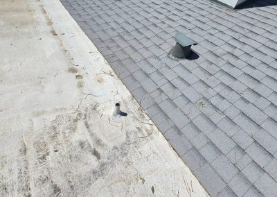 A close-up view of a rooftop showing a transition between a flat, weathered white roof surface and a section with gray asphalt shingles. Two vent pipes protrude from the roof near the transition line.