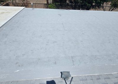 A flat, gray rooftop with a few visible seams and some scattered marks. A small portion of a white roof section is seen on the left, and tree branches are visible in the background. A plastic chair is at the roof’s edge.