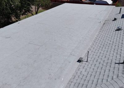 A flat, light gray roof next to a sloped shingle roof with several vents and pipes; trees and neighboring buildings are visible in the background.