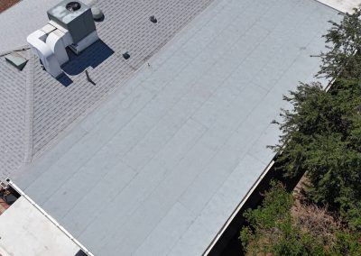 Aerial view of a building roof with two distinct sections: one with gray shingles and ventilation units, the other with a flat, smooth gray surface. Trees partially surround the edge of the structure.