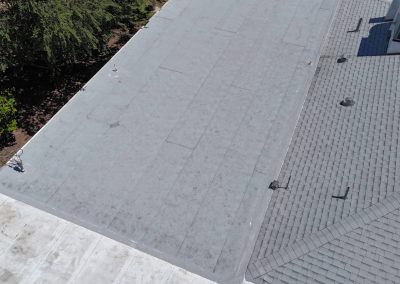 Aerial view of a building's roof, showing a flat, gray, asphalt section next to a sloped, shingled section. Trees and grass are visible on the left side of the image.