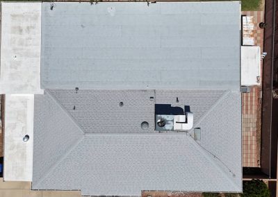 Aerial view of a house roof with both flat and pitched sections, various vents and equipment, surrounded by paved and landscaped areas, fences, and some vegetation.
