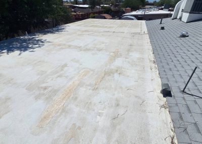 A flat, light-colored roof with worn and stained areas is next to a sloped, gray-shingled roof under a sunny sky, with some vents and pipes visible on both roofs. Trees and houses are in the background.