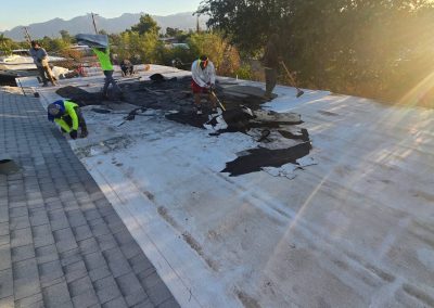 Several workers repair a flat, white roof, removing worn black roofing material under bright sunlight. Nearby trees, rooftops, and distant mountains are visible in the background.