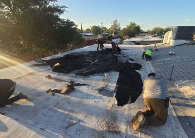Several workers are repairing a large, damaged section of a flat white roof under early morning sunlight, with trees and buildings visible in the background.