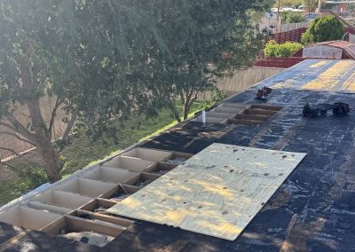 A rooftop under repair with exposed wooden beams, a sheet of plywood partially covering the opening, and tools scattered nearby. Trees and a fence are visible beside and below the roof.