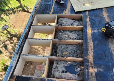 A roof section with decking removed, exposing insulation and joists beneath. Tools and a tool belt are on the roof nearby. Trees, grass, and a birdbath are visible on the ground below.