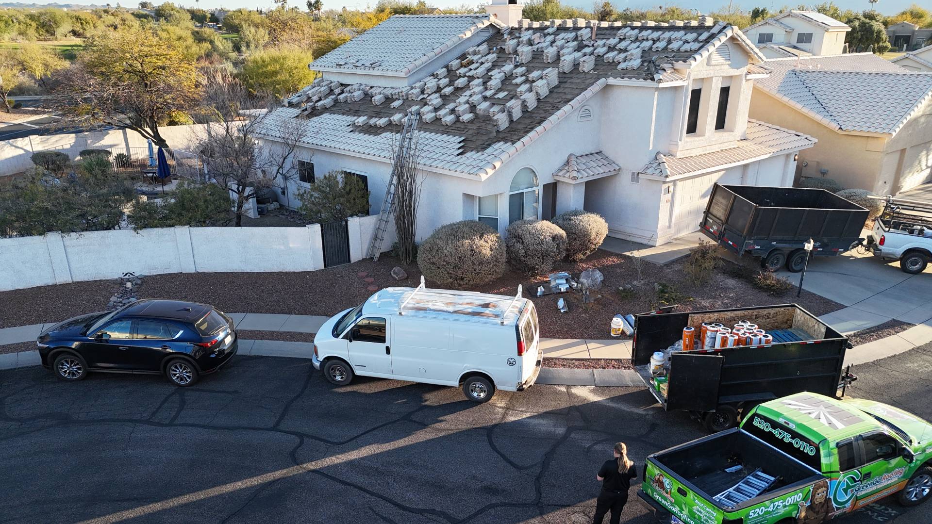 Aerial view of a house with roof tiles removed, construction materials on the driveway, a white van, a black SUV, a trailer, and a green truck parked on the street, indicating ongoing roof repair work.