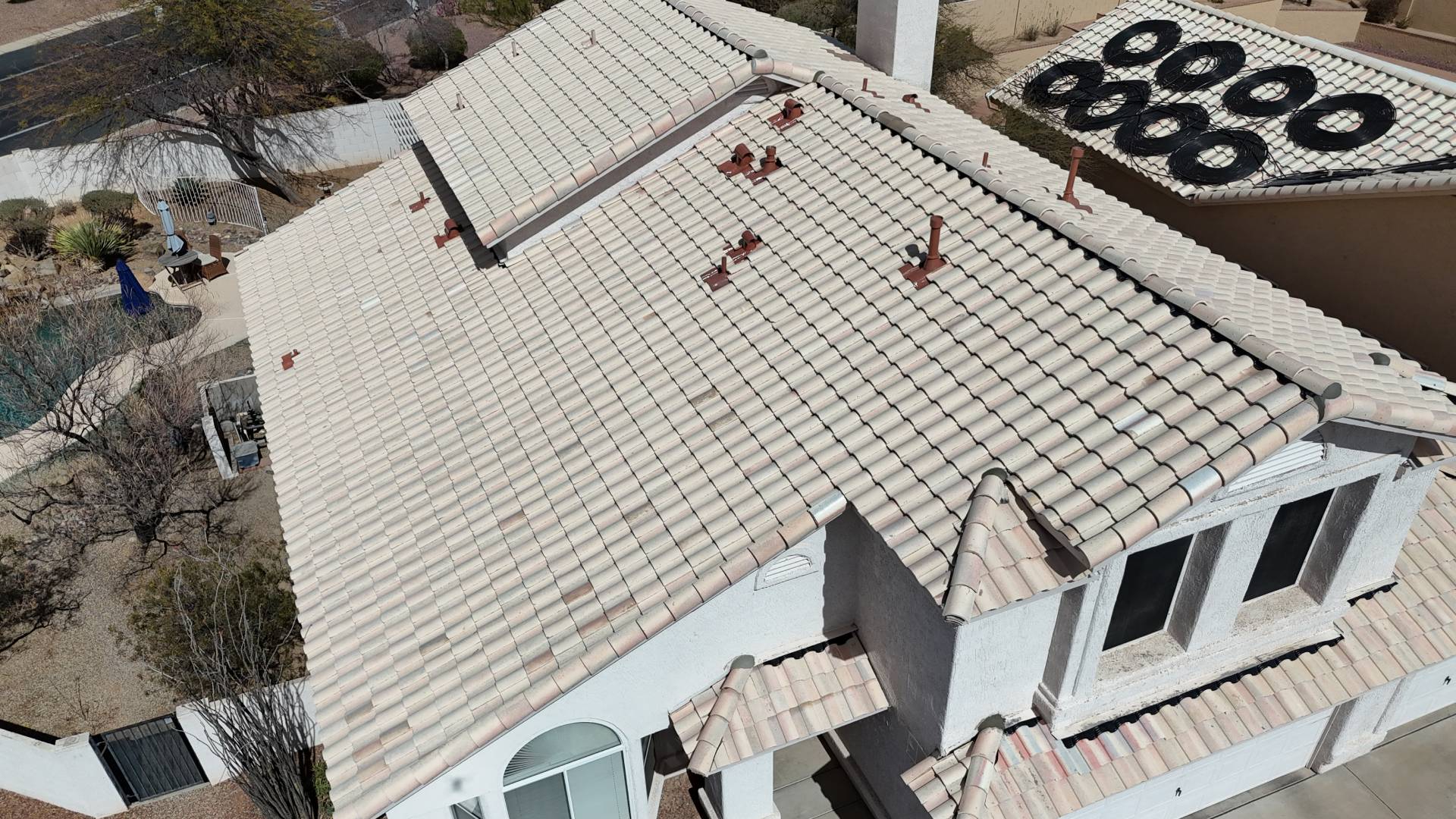 Aerial view of a large house with a light-colored tiled roof, showing multiple vent pipes. The house is surrounded by a dry landscape with sparse vegetation. Neighboring roof has several black circular objects.