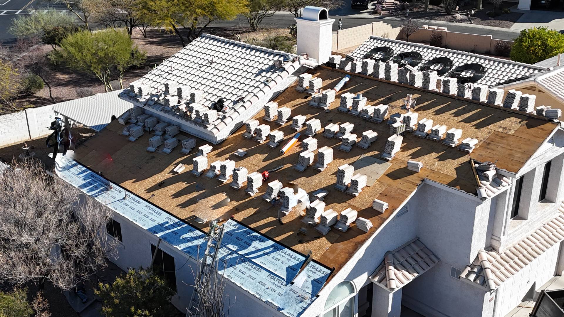 Aerial view of a house with workers on the roof preparing for new tiles. Bundles of roofing tiles are evenly spaced across the wooden roof, and some sections are covered with blue underlayment. Trees and roads surround the house.