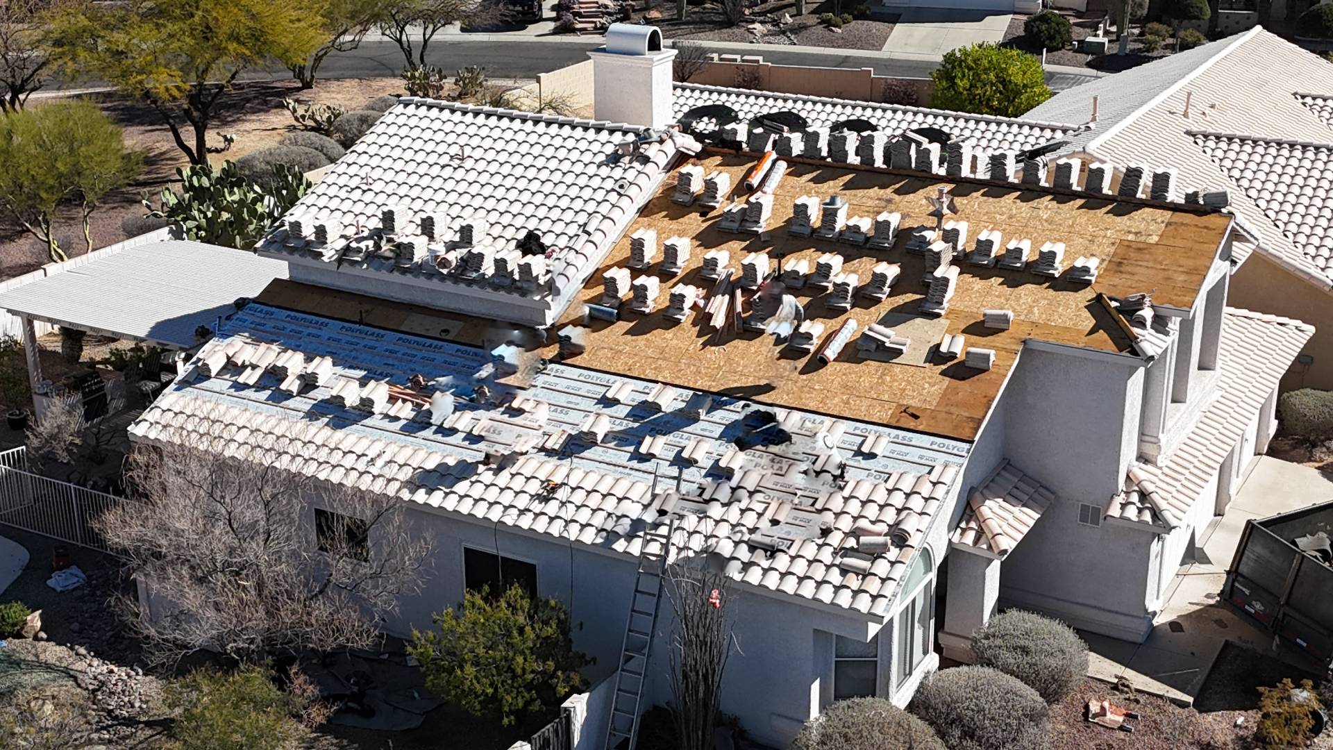 An aerial view of a house with workers repairing the roof; white tiles have been removed from part of the roof, exposing wooden boards, and new materials and tiles are stacked and spread across the surface.