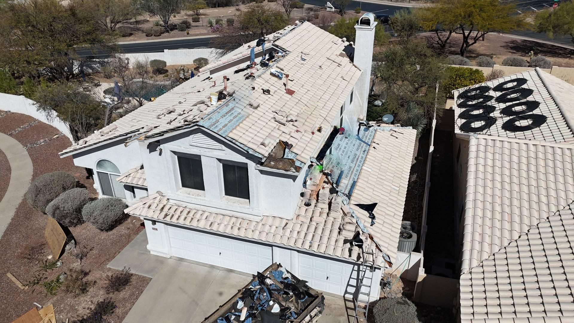 Aerial view of a house with workers repairing or installing a new roof. Tools, materials, and old roofing debris are scattered on the roof and driveway. Neighboring houses and desert landscaping are visible nearby.