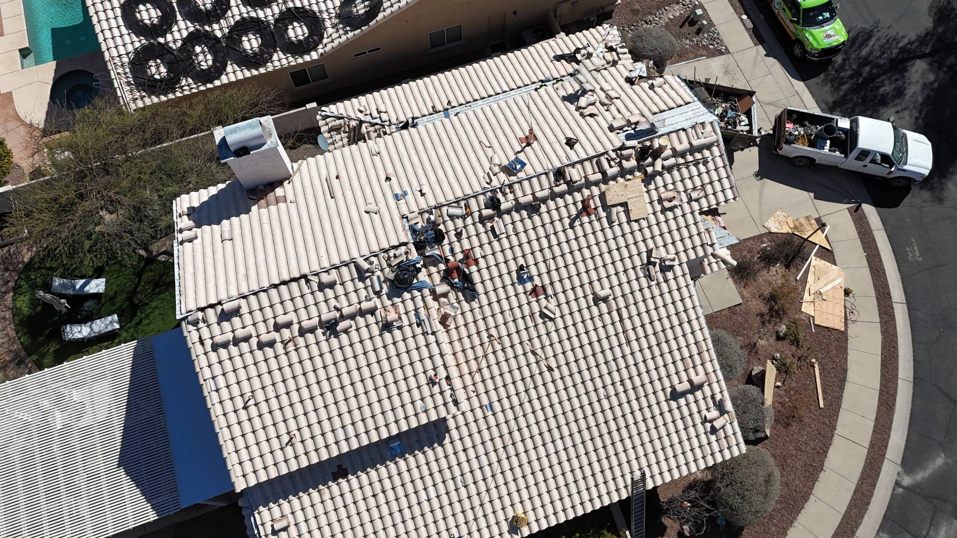 Aerial view of workers repairing or installing a tile roof on a house. Several vehicles and construction materials are visible on the driveway and street, with tools and tiles spread across the rooftop.