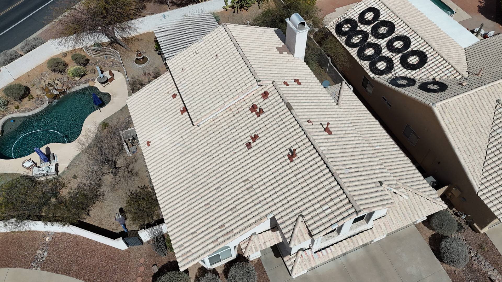 Aerial view of a beige-tiled rooftop on a suburban house, with a backyard pool, desert landscaping, and neighboring homes visible. Circular objects are on the adjacent roof. Sidewalks and a driveway are also shown.