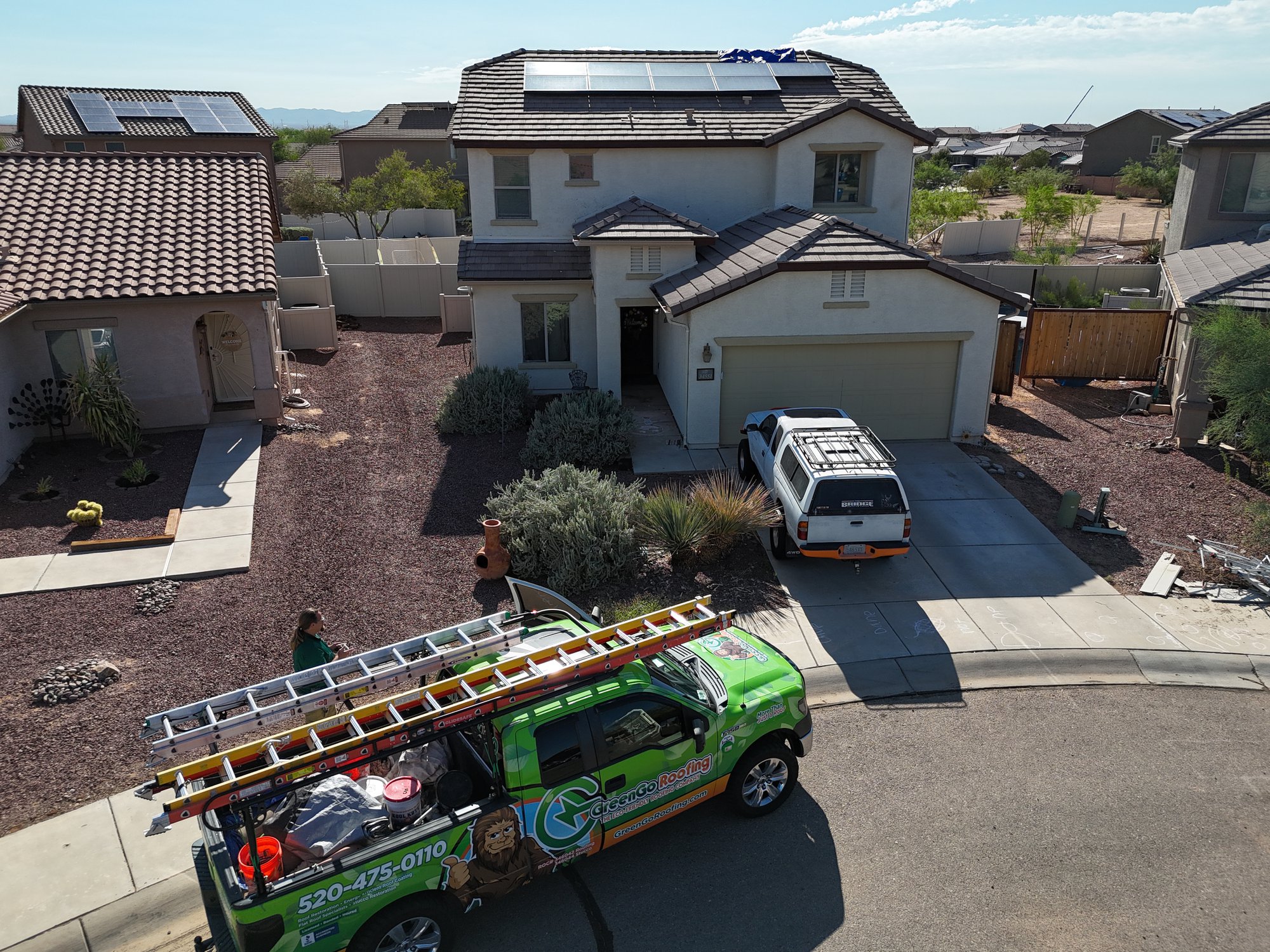 A two-story house with solar panels on the roof is seen from above. A green service truck and a white car are parked in the driveway. Two people walk on the sidewalk toward the house. The yard has desert landscaping. default