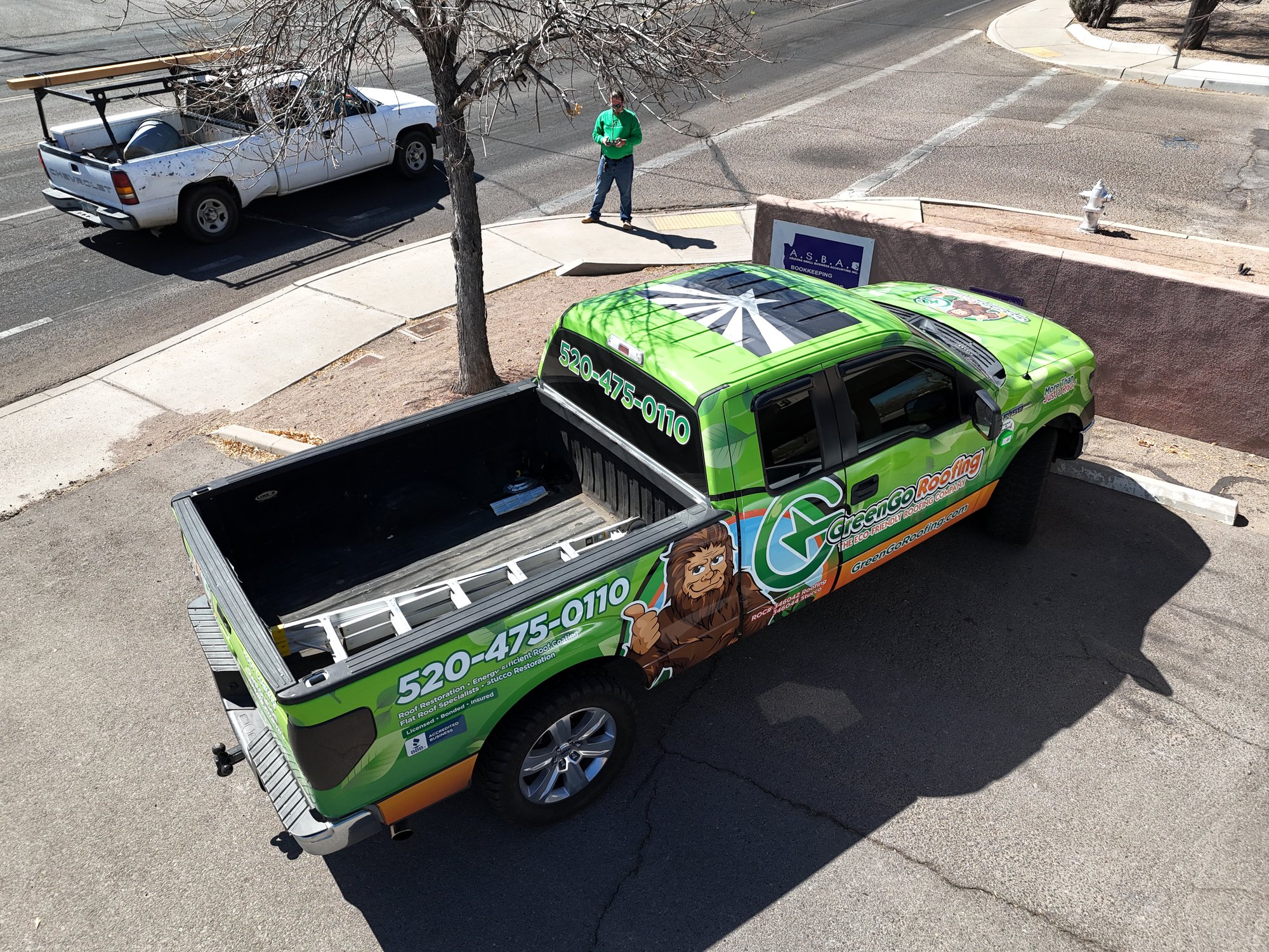 A green pickup truck with business advertising and a cartoon lion on the side is parked on a street. A man in a green shirt stands nearby, and another white truck is visible across the street. default