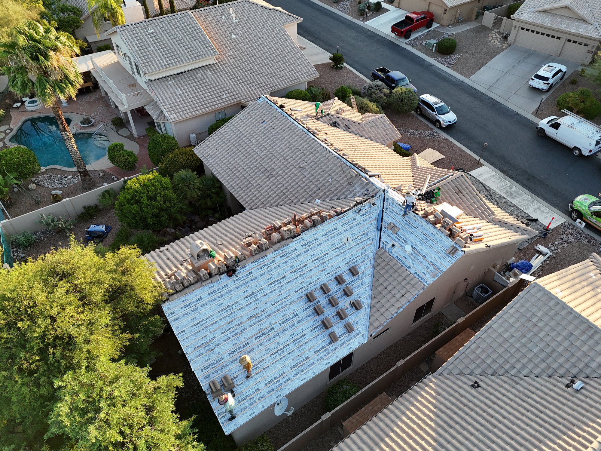 Aerial view of a house with workers installing roofing materials on a partially covered roof, surrounded by neighboring homes, trees, a pool, and vehicles parked along the street. default