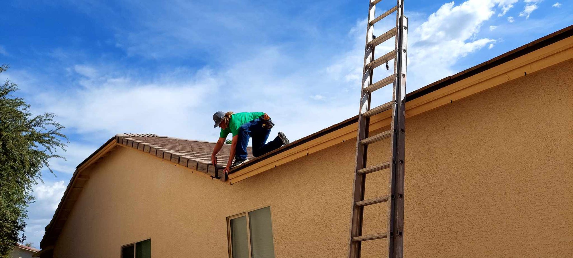 A person wearing a hat and green shirt works on the roof of a beige house under a blue sky, with a tall ladder leaning against the side of the building. 8 Emergency Roof Repair Tips for Tucson Residents cover