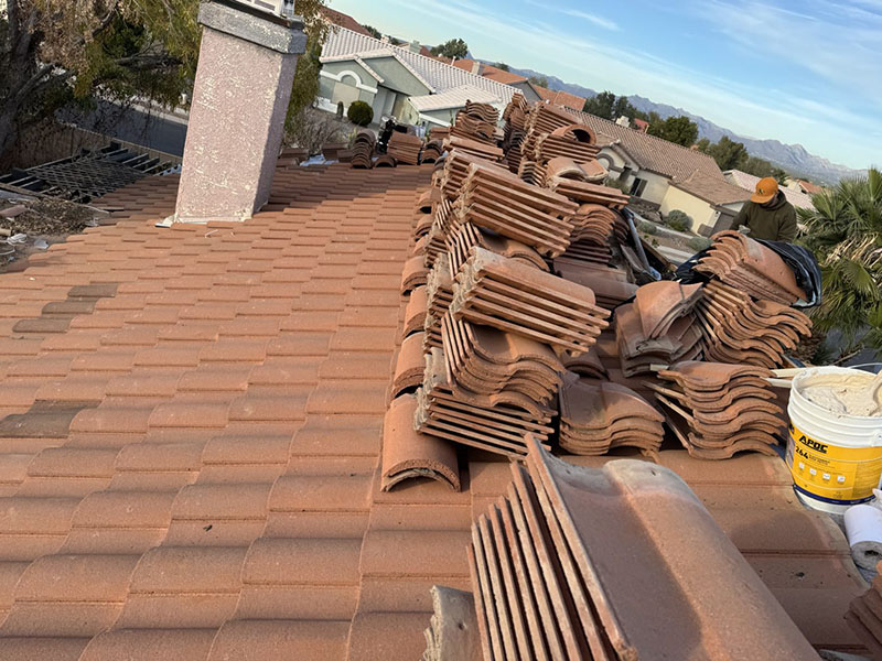 Stacks of reddish-brown roof tiles are arranged on a sloped rooftop. A person works in the background near buckets and tools. Houses, trees, and distant mountains are visible under a partly cloudy sky.